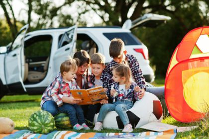 Family spending time together. Mother reading book outdoor with kids against their suv car.