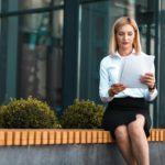 Female office worker sits on a bench near building and looks at documents