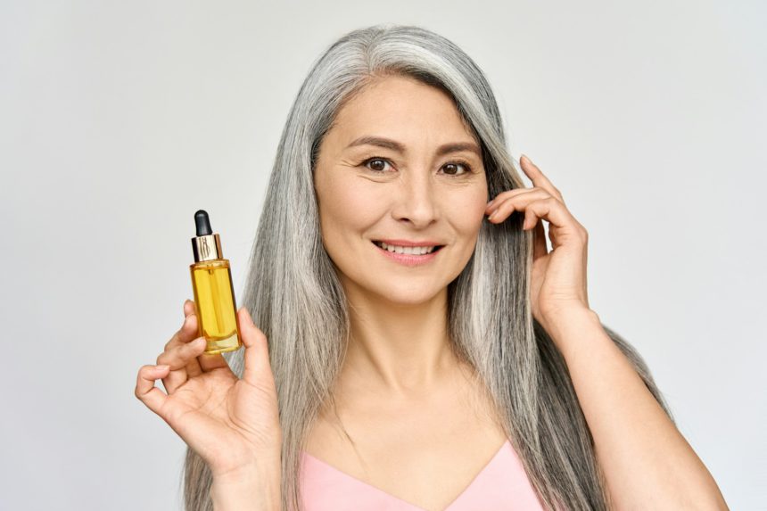 Headshot of smiling mid age Asian woman holding hair and skin care oil serum.