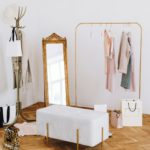 Rack with stylish women's clothes, grey pouf and mirror indoors. Interior design