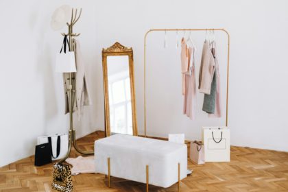 Rack with stylish women's clothes, grey pouf and mirror indoors. Interior design