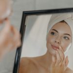 Woman wears minimal makeup, takes care of complexion and lips, looks at herself in mirror