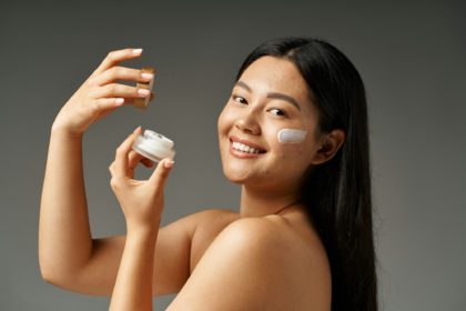 young asian woman with acne prone skin holding container with cream on grey background, skin issue